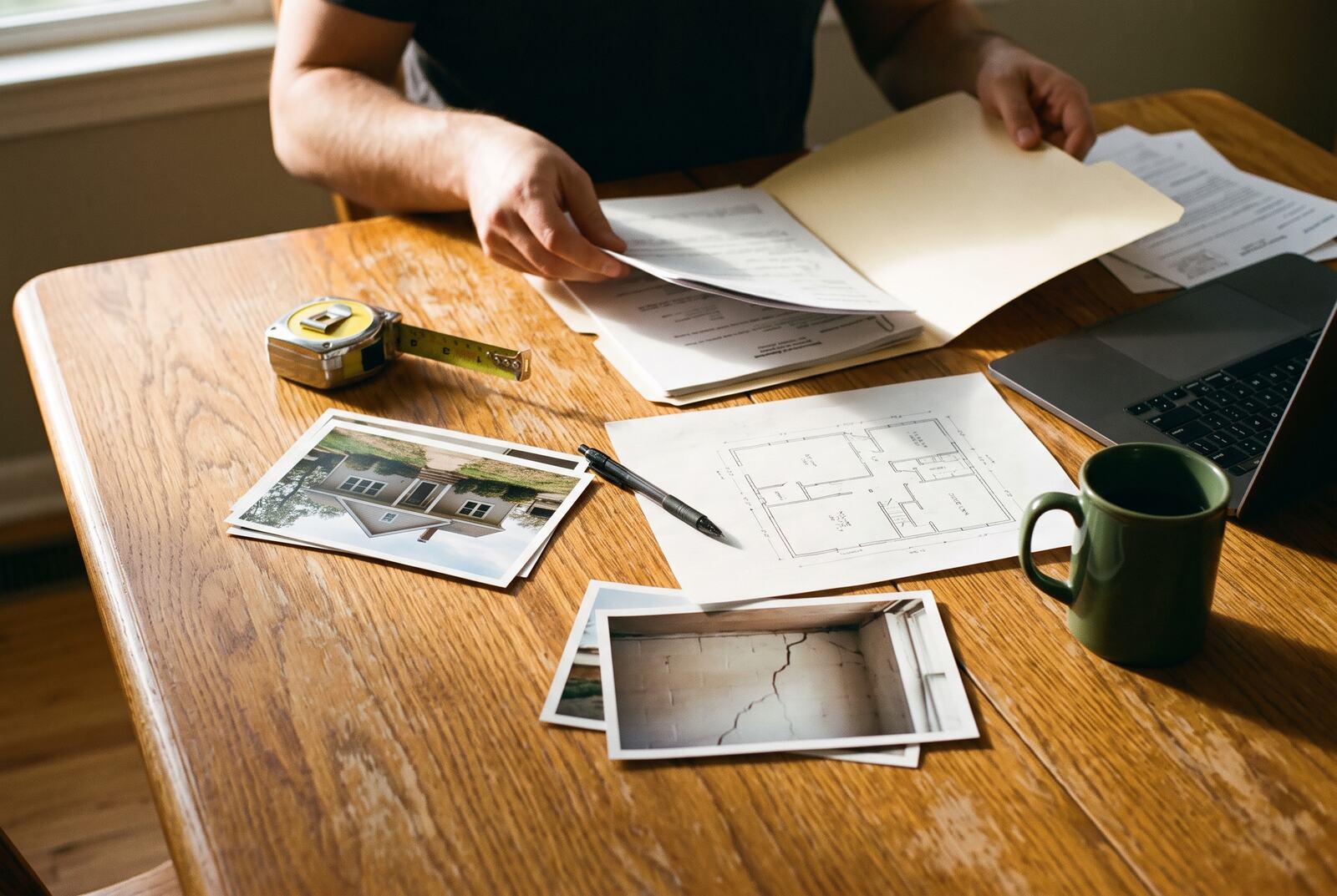 Hands sorting printed photos of a house, a folder of papers and a tape measure on a kitchen table